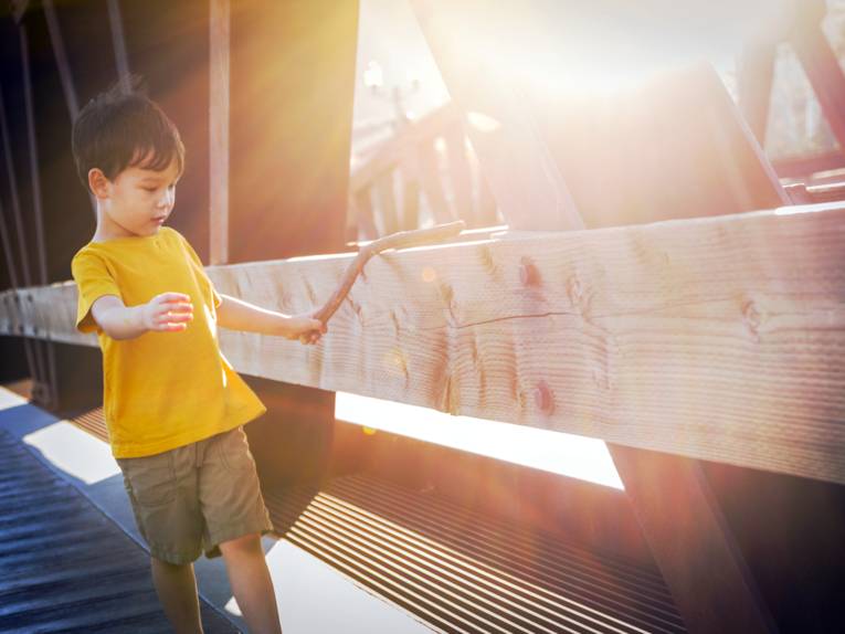 Ein Junge hat in der linken Hand einen kräftigen, kurzen, krummen Stock und führt ihn über ein Brückengeländer aus Holz. Die Sonne tauscht die Szene in warmes Licht.