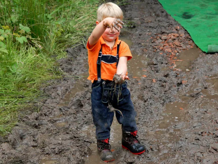 Ein kleiner, blonder Junge trägt eine Latzhose, ein orangefarbenes Polohemd und Schnürstiefel. Er streckt die Arme aus und lässt Matsch von der oberen Hand in die untere fallen. Er steht auf durchnässtem, matschigem Boden. Am Rand wächst hohes Gras. 