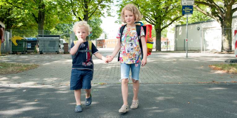 Ein Junge und ein Mädchen reichen sich die Hände und überqueren eine Straße. Kedes Kind trägt einen Schulranzen.