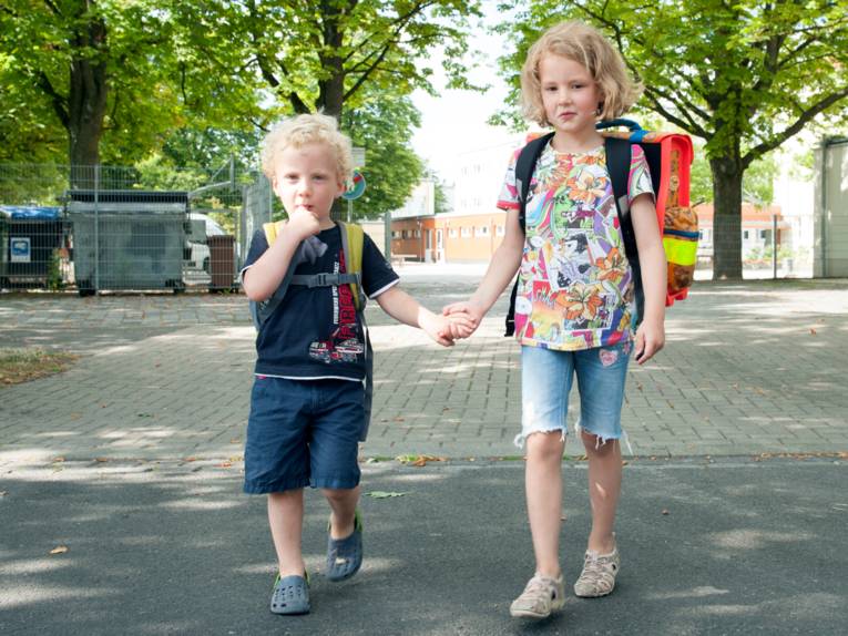Ein Junge und ein Mädchen reichen sich die Hände und überqueren eine Straße. Kedes Kind trägt einen Schulranzen.
