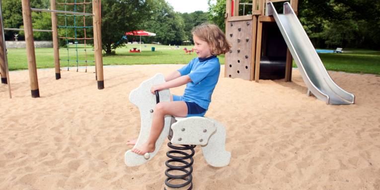 Ein Kind reitet auf einem Schaukelpferd, im Hintergrund sind eine Rutsche und ein Klettergerüst.