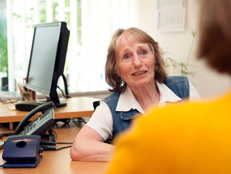 Eine ältere Frau sitzt in an einem Tisch, im Hintergrund sind ein Telefon und ein PC-Monitor zu sehen. Im Vordergrund ist unscharf der Rücken und der Kopf einer jüngeren Frau zu erkennen. Beide schauen sich an und sprechen miteinander.
