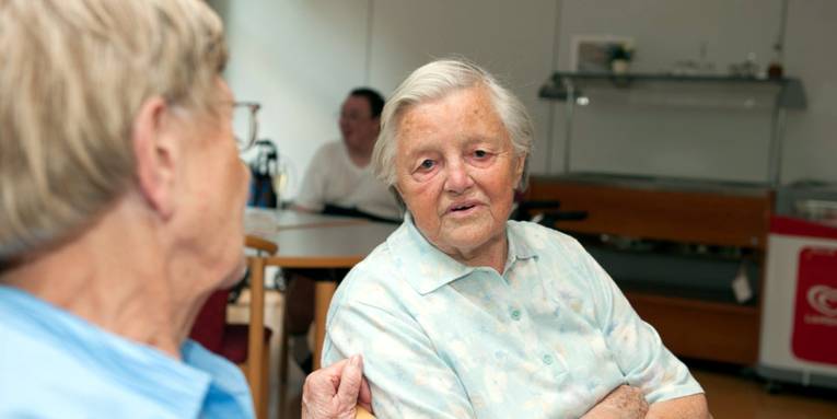 Zwei ältere Menschen sitzen nebeneinander auf Stühlen und sprechen miteinander.