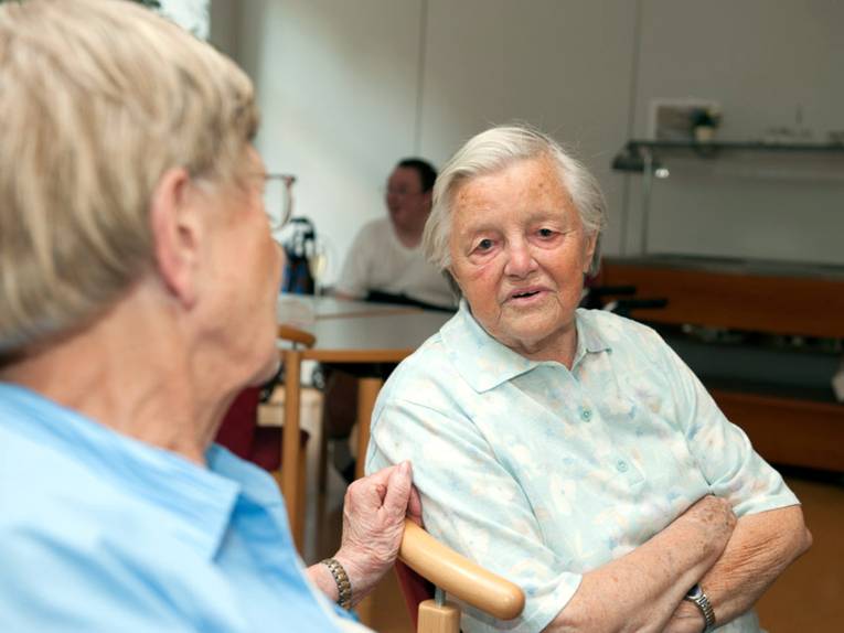 Zwei ältere Menschen sitzen nebeneinander auf Stühlen und sprechen miteinander.