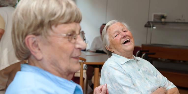 Zwei ältere Menschen sitzen nebeneinander auf Stühlen und lachen.
