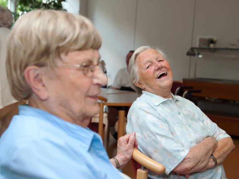 Zwei ältere Menschen sitzen nebeneinander auf Stühlen und lachen.