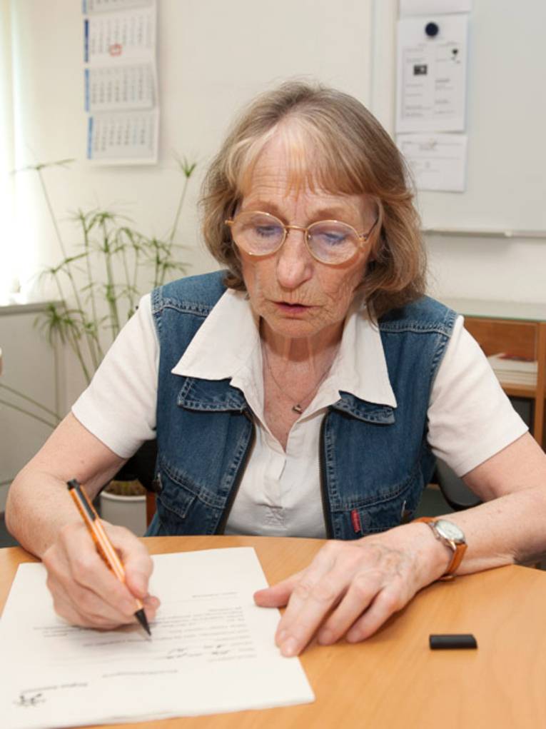 Eine ältere Frau sitzt an einem Tisch und füllt mit einem Stift ein Formular aus.