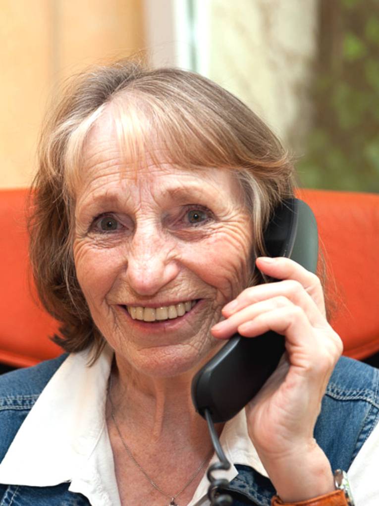 Eine ältere Dame sitzt auf einem Bürostuhl, hält einen Telefonhörer in ihrer linken Hand und lächelt in die Kamera.