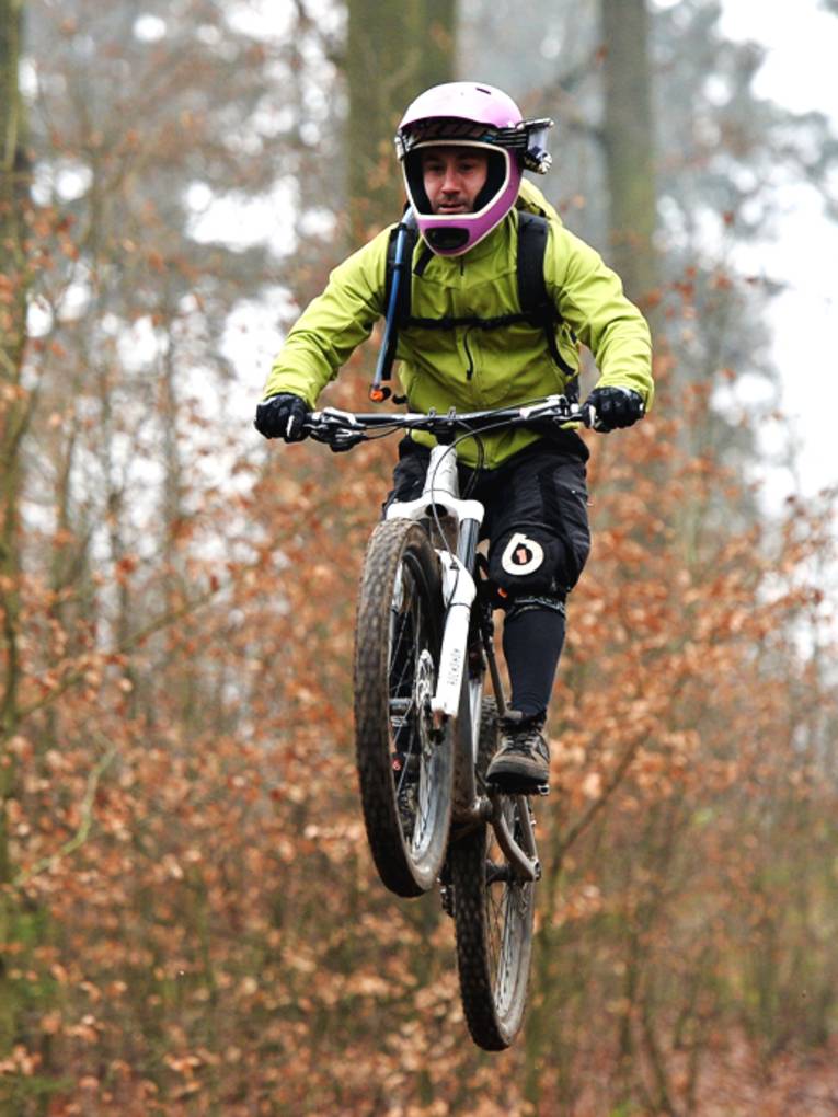 Ein Mointainbiker trägt eine hellgrüne Jacke und einen rosafarbenen Helm. Er fliegt gerade samt Fahrrad nach einem Sprung über dem Boden. Am linken Rand steht ein älterer Herr mit einem Jungen, beide schauen zu.