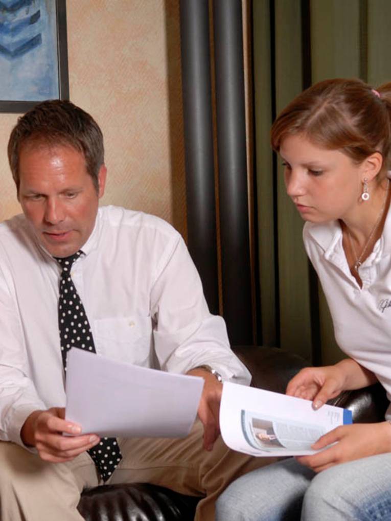Eine Frau schaut auf die Hände eines Mannes, er zeigt etwas auf einem Papier. Die Szene findet in einer Sitzecke mit Ledersesseln statt.