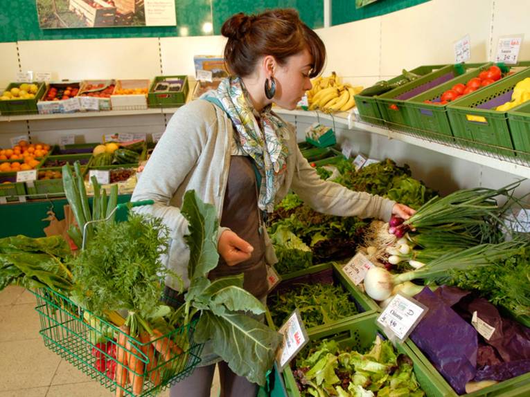 Eine junge Frau im Supermarkt. Sie ist gerade in der Obst- und Gemüseabteilung. In ihrem Einkaufskorb befindet sich bereits einiges an Gemüse (unter anderem ein Bund Karotten) und die Frau wählt weiteres Gemüse aus. In der linken Hand hat sie ein Bund rote Frühlingszwiebeln.