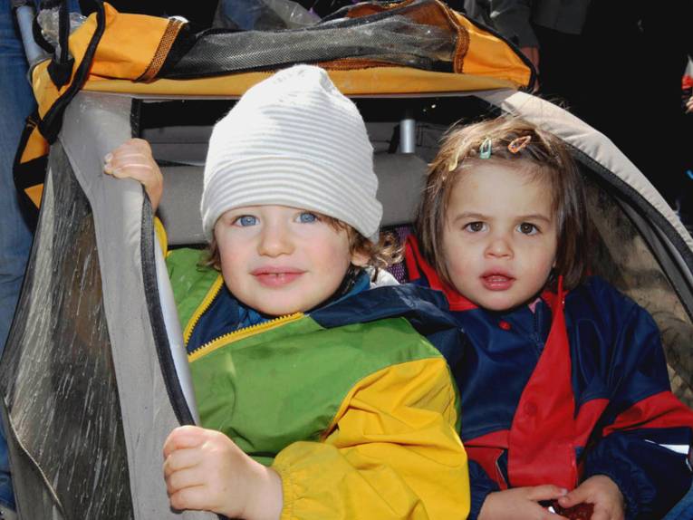 Ein Junge und ein Mädchen sitzen nebeneinander in einem Fahrradanhänger. Bunte Regenkleidung schützt sie vor Wind und Wetter.