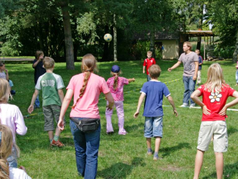 Auf dem Bild sind zehn Kinder, eine Betreuerin und ein Betreuer abgebildet, die gemeinsam mit einem Ball spielen. Folienband auf dem Rasen markiert das Spielfeld.
