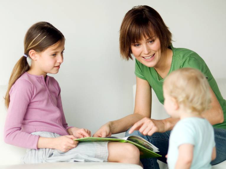 Eine erwachsense Frau schaut sich gemeinsam mit einem Mädchen ein Bilderbuch an, sie sitzen dabei. Ein Kleindkind steht vor den beiden Sitzenden.