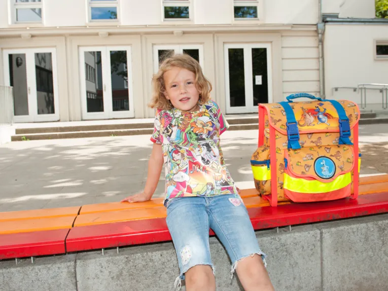 Mädchen mit Schulranzen auf der Bank vor einem älteren Gebäude sitzend