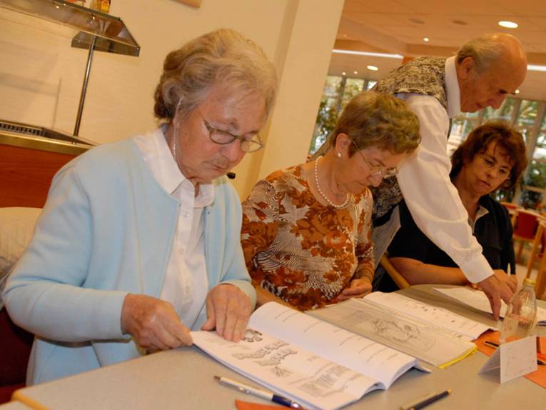 Drei Frauen sitzen und ein Mann steht an einem Tisch. Die beiden ältteren Frauen blättern in Unterlagen und der Männer zeigt der jüngeren Frau ezwas auf einem Blatt.