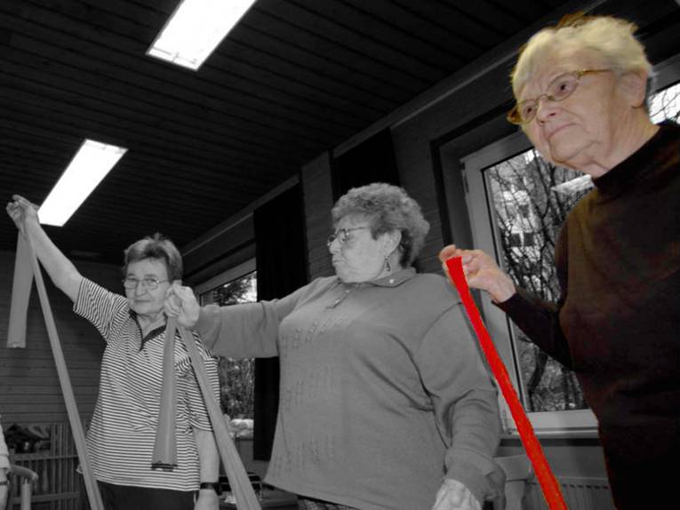 Drei ältere Damen trainieren ihre Muskeln mit Therapiebändern. Das Bild ist auf die Farben Schwarz und Weiß reduziert, nur die älteste Dame (rechts) setzt sich farbig ab.