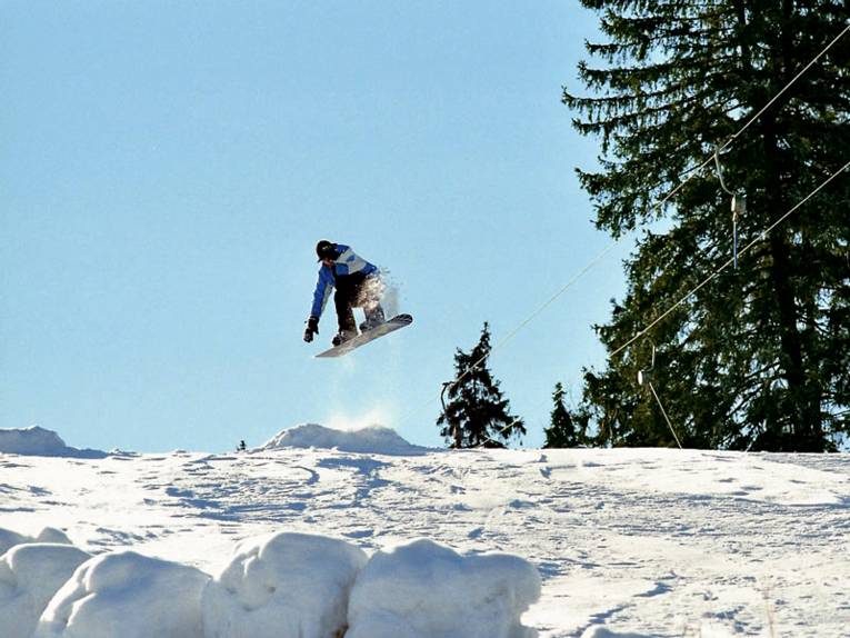 Nach dem Sprung über eine kleine Schanze fliegt ein Snowboarder über eine weiße Winterlandschaft. Der Himmel über ihm strahlt in hellem Blau.