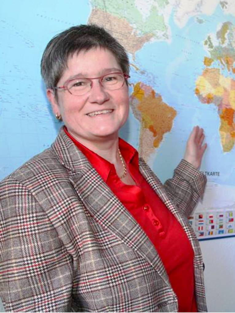 Porträtaufnahme einer Frau mit kurzen Haaren und Brille, die mit der Hand auf eine Weltkarte zeigt.