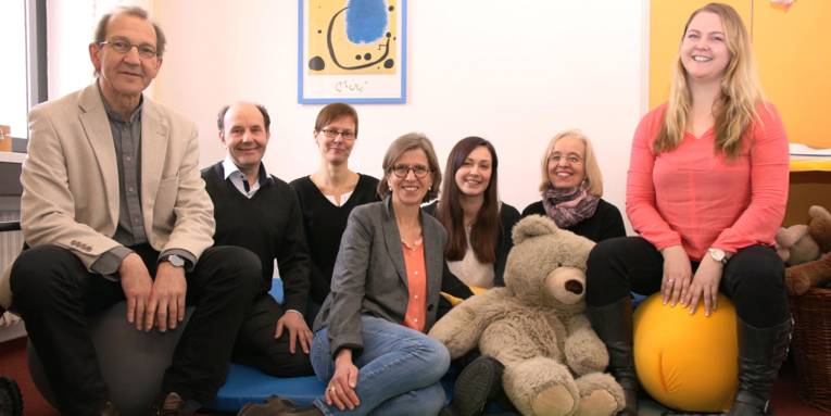 Ein Team bestehend, aus fünf Frauen und zwei Männern, in einem Raum. Fünf Personen sitzen mit einem großen Teddybären auf einer Matte auf dem Boden. Rechts und links von ihnen sitzen ein Mann und eine junge Frau jeweils auf einem Ball.