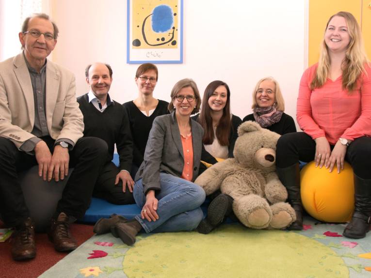 Ein Team bestehend, aus fünf Frauen und zwei Männern, in einem Raum. Fünf Personen sitzen mit einem großen Teddybären auf einer Matte auf dem Boden. Rechts und links von ihnen sitzen ein Mann und eine junge Frau jeweils auf einem Ball.
