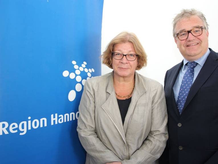 Hauke Jagau und Andrea Fischer stehen in einem Raum mit weißer Wand. Links neben ihnen ist das Logo der Region Hannover in weiß auf blauem Hintergrund zu sehen.