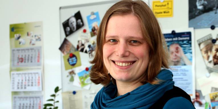 Portraitaufnahme einer Frau, hinter ihr sind eine weiße Magnettafel, ein Kalender und eine Grünpflanze.