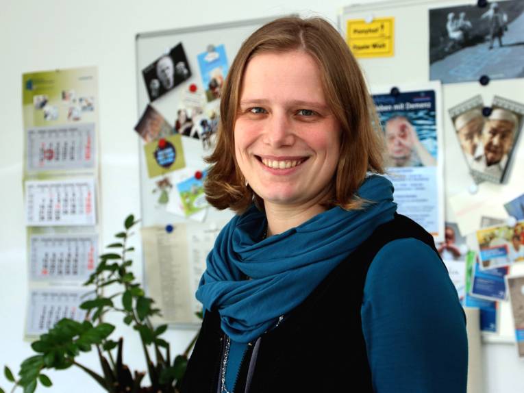 Portraitaufnahme einer Frau, hinter ihr sind eine weiße Magnettafel, ein Kalender und eine Grünpflanze.