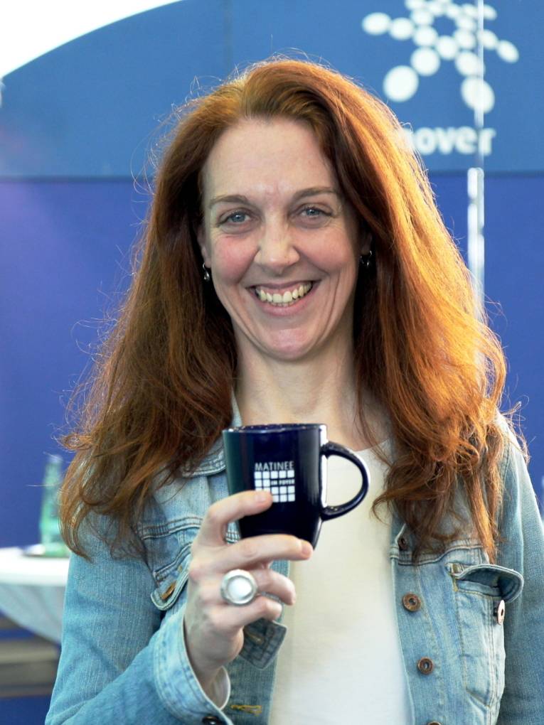Eine Frau mit langen, roten Haaren steht lächelnd vor zwei Stehtischen und einer blauen Ausstellungswand. In der rechten Hand hält sie eine Tasse mit dem Logo der Matinee im Foyer auf Brusthöhe vor sich.