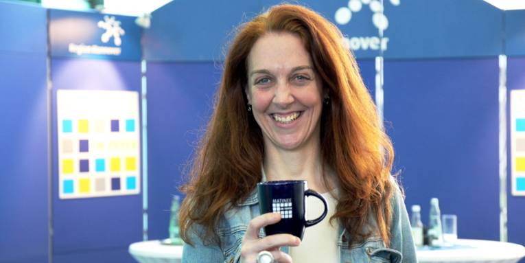 Eine Frau mit langen, roten Haaren steht lächelnd vor zwei Stehtischen und einer blauen Ausstellungswand. In der rechten Hand hält sie eine Tasse mit dem Logo der Matinee im Foyer auf Brusthöhe vor sich.