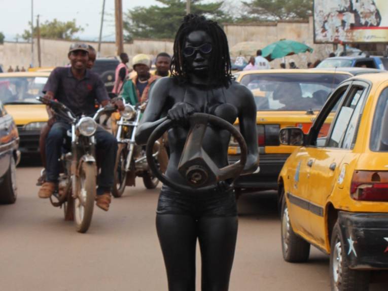 Eine mit Körperfarben schwarz angemalte Frau, die ein Autolenkrad in der Hand hält. Im Hintergrund Autos und Menschen auf Motorrollern 