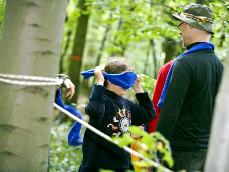 Ein Kind hat die Augen verbunden und steht neben einem Kletterhindernis. Davor steht ein Mann mit Rangerhut.
