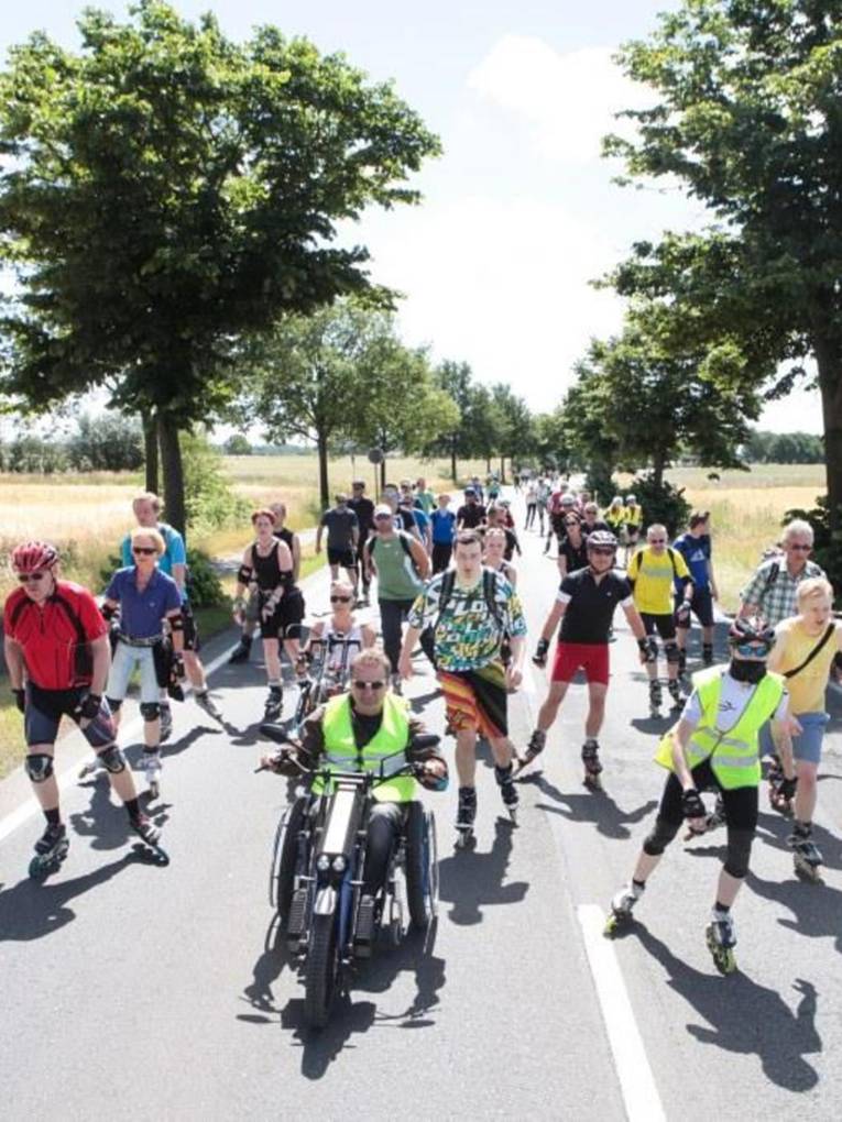 Eine große Gruppe aus Skatern fährt über eine Landstraße, ein Handradfahrer fährt vorne mit.