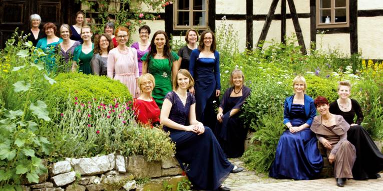 Frauen tragen Abendkleider und stehen oder sitzen in einem Garten.