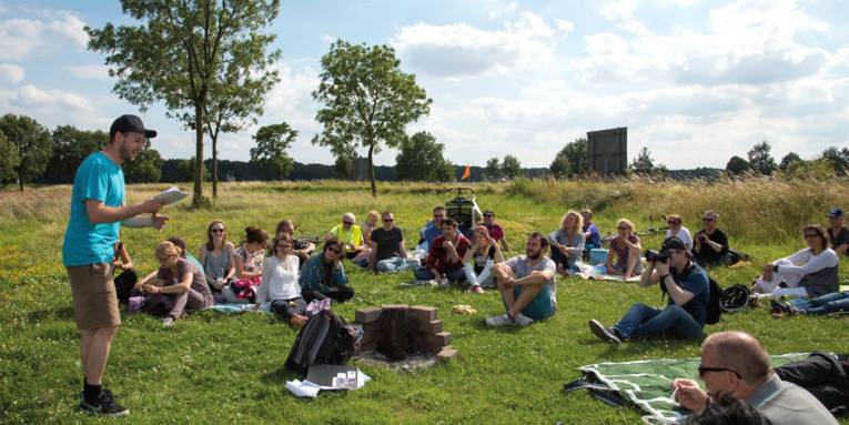 Menschen sitzen auf einer Wiese, davor steht ein Mann und trägt einen Text vor.