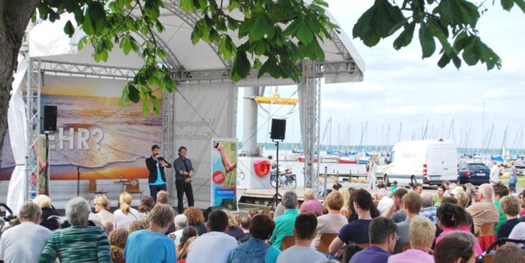 Zwei Männer stehen auf einer schwimmenden Bühne, Publikum verfolgt das Programm.