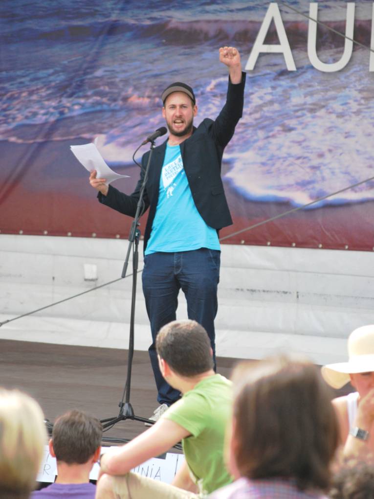 Ein Mann steht an einem Mikrofon und hebt die linke Faust, rechts hält er mehrere Blatt Papier.