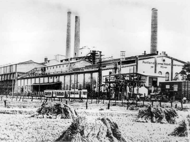 Historische Aufnahme einer Cementfabrik, im Vordergrund Getreidegarben und eine Straßenbahn