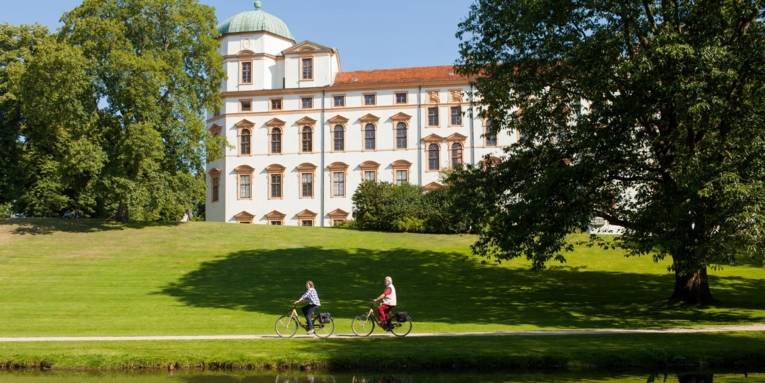 Im Vordergrund ein Teich, dahinter ein Grünstreifen mit zwei Radfahrern und im Hintergrund ein Schloss.