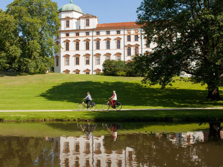 Im Vordergrund ein Teich, dahinter ein Grünstreifen mit Radfahrern und im Hintergrund ein Schloss, an einem sonnigen Tag mit blauem Himmel