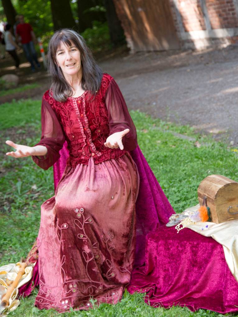 Eine Frau trägt ein rotes Gewand und sitzt neben einer kleinen Schatztruhe.