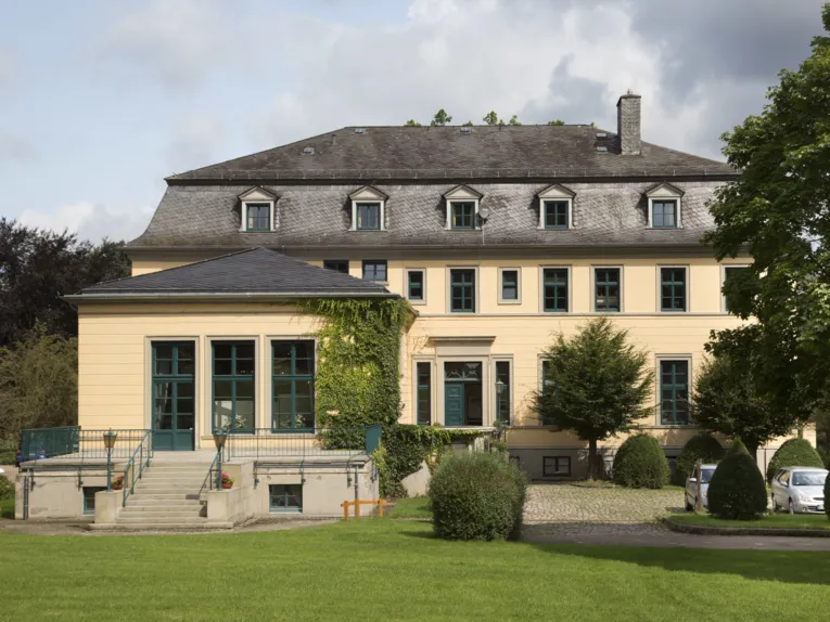 Blick aus einem Park auf ein Gebäude mit Freitreppe und Auffahrt