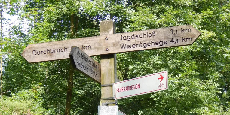 Ein Wegweiser aus Holz in einem Wald, auf dem unter anderem "Durchbruch", "Jagdschloss" und "Wisentgehege" mit Kilometerangabe ausgewiesen sind.