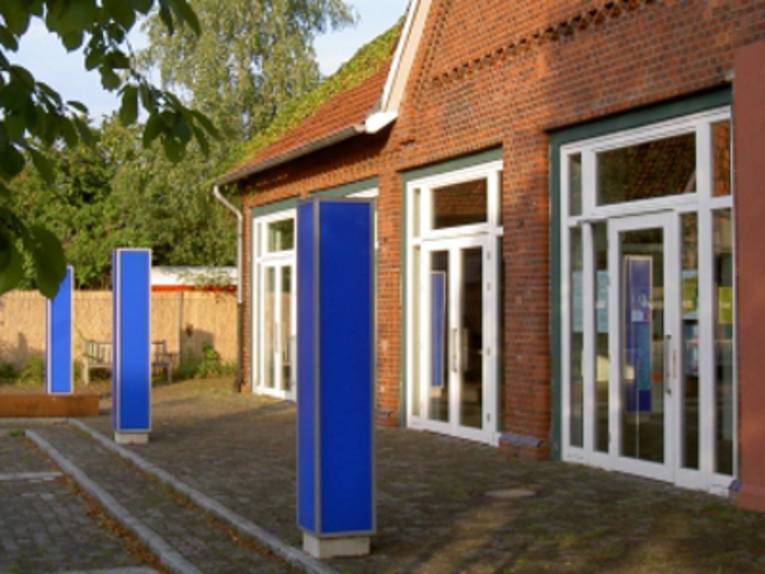 Vor einem roten Backsteinbau stehen drei blaue Säulen.