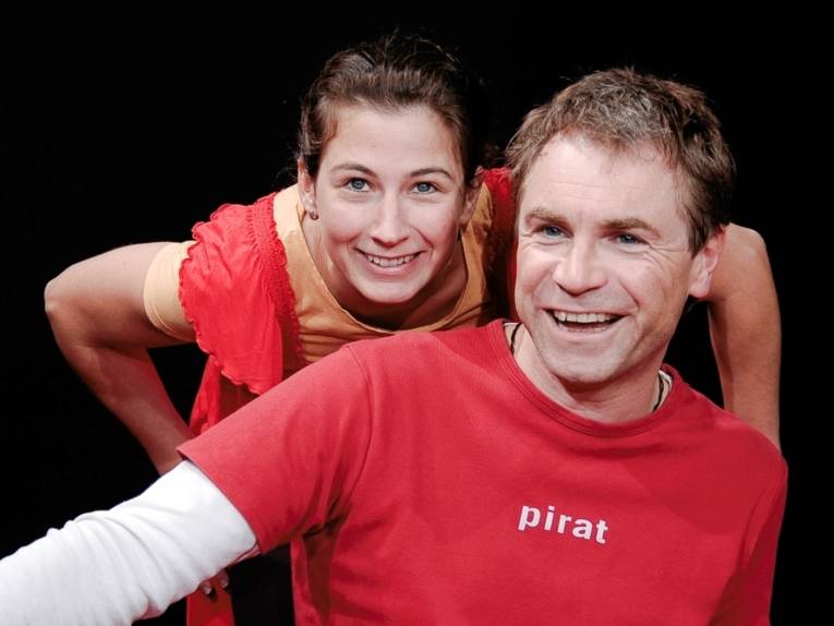 Zwei Schauspieler vor schwarzem Hintergrund. Rechts ein Mann in rotem T-Shirt mit Aufdruck "pirat", dahinter eine Frau in einem roten Kleidung und gelben T-Shirt.