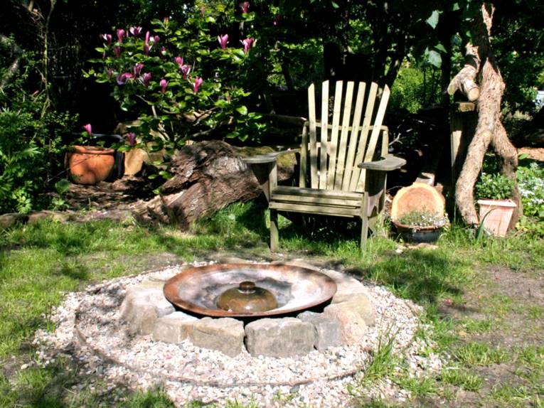 Ein Holzstuhl steht in einem Garten, im Hintergrund blüht Kirschlorbeer. Vor dem Stuhl steht eine Metallschale, die von Feld- und Kieselsteinen eingefasst ist.