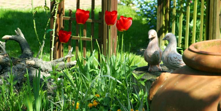 In einem Beet wachsen verschiedene Pflanzen, unter anderem rote Tulpen. Auf einem abgesägten Baumstamm stehen Tonfiguren in Taubenform.