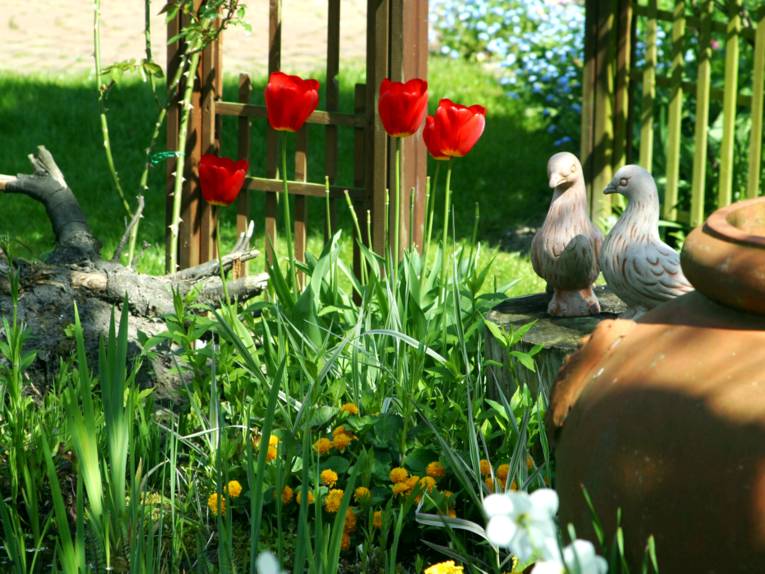 In einem Beet wachsen verschiedene Pflanzen, unter anderem rote Tulpen. Auf einem abgesägten Baumstamm stehen Tonfiguren in Taubenform.