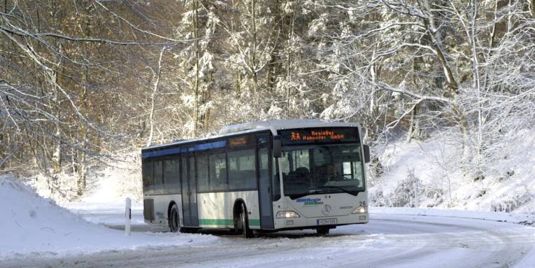 Ein Bus der RegioBus GmbH fährt auf einer verschneiten Straße um die Kurve, es geht bergauf. Neben der Straße stehen schneebedeckte Bäume.