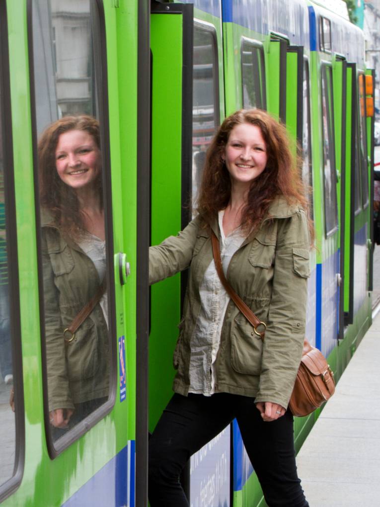 Eine junge Frau steigt in eine Stadtbahn der üstra, ihr Gesicht wird in der Seitenscheibe gespiegelt. Am Fahrzeugkopf steigt eine weitere Person ein.